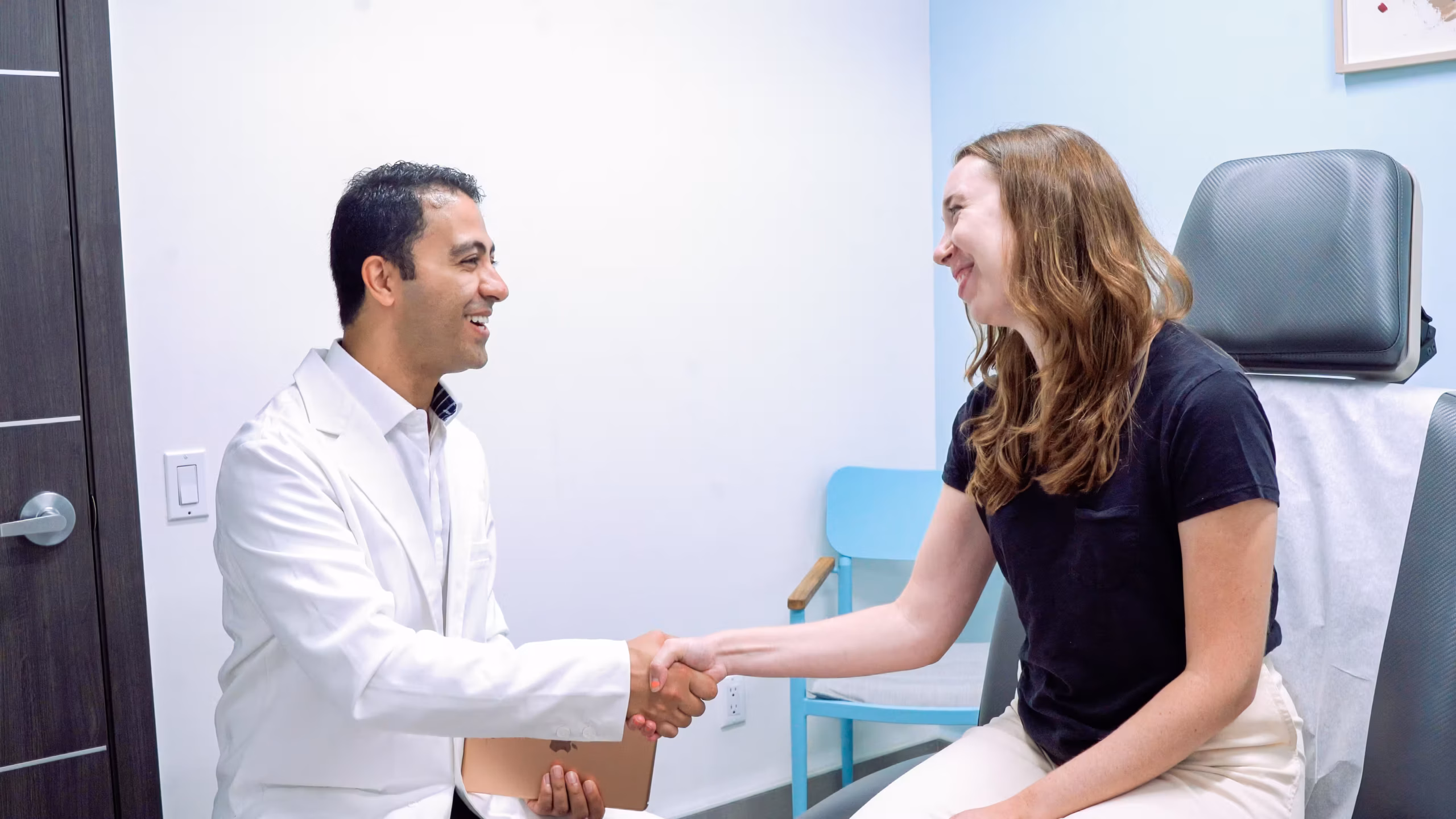 side-view of a doctor, wearing a white lab coat and a collared shirt, smiling while shaking the hand of a female patient. The doctor is on the left, looking toward the patient. The patient is on the right, wearing a black t-shirt and light-colored pants, sitting on an examination table, and smiling back at the doctor.